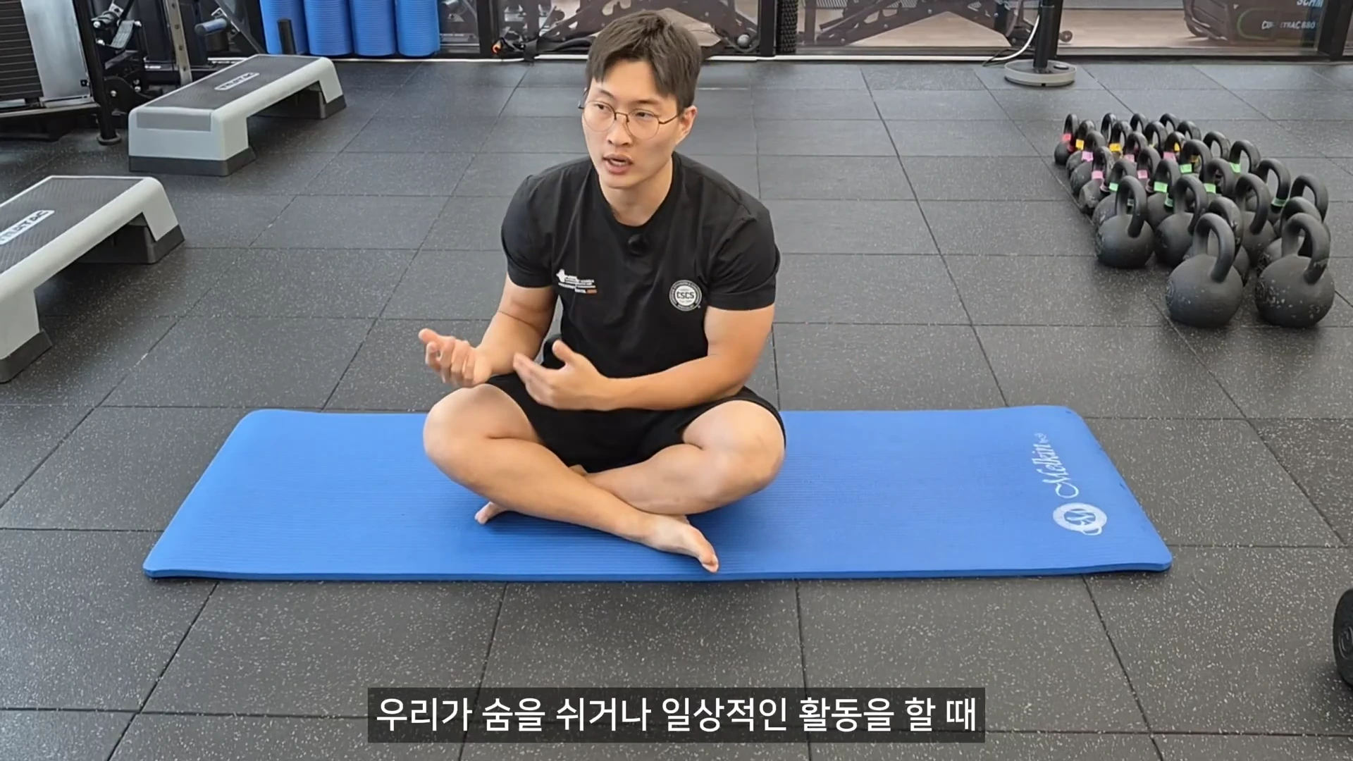 운동복을 입은 안경 쓴 남성이 헬스장 바닥에 놓인 파란색 요가 매트 위에 가부좌를 틀고 앉아 손동작을 곁들여 설명하고 있다.
