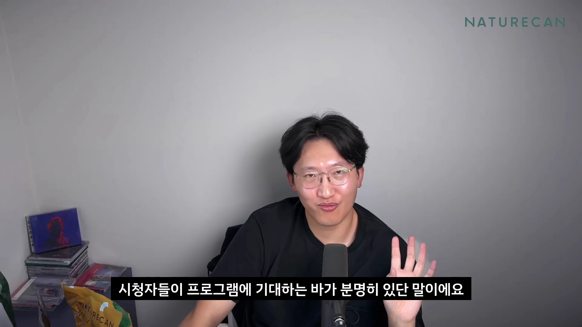 안경을 쓴 남성이 검은색 티셔츠를 입고 마이크 앞에 앉아 손을 들어 제스처를 하며 이야기를 나누고 있다.
