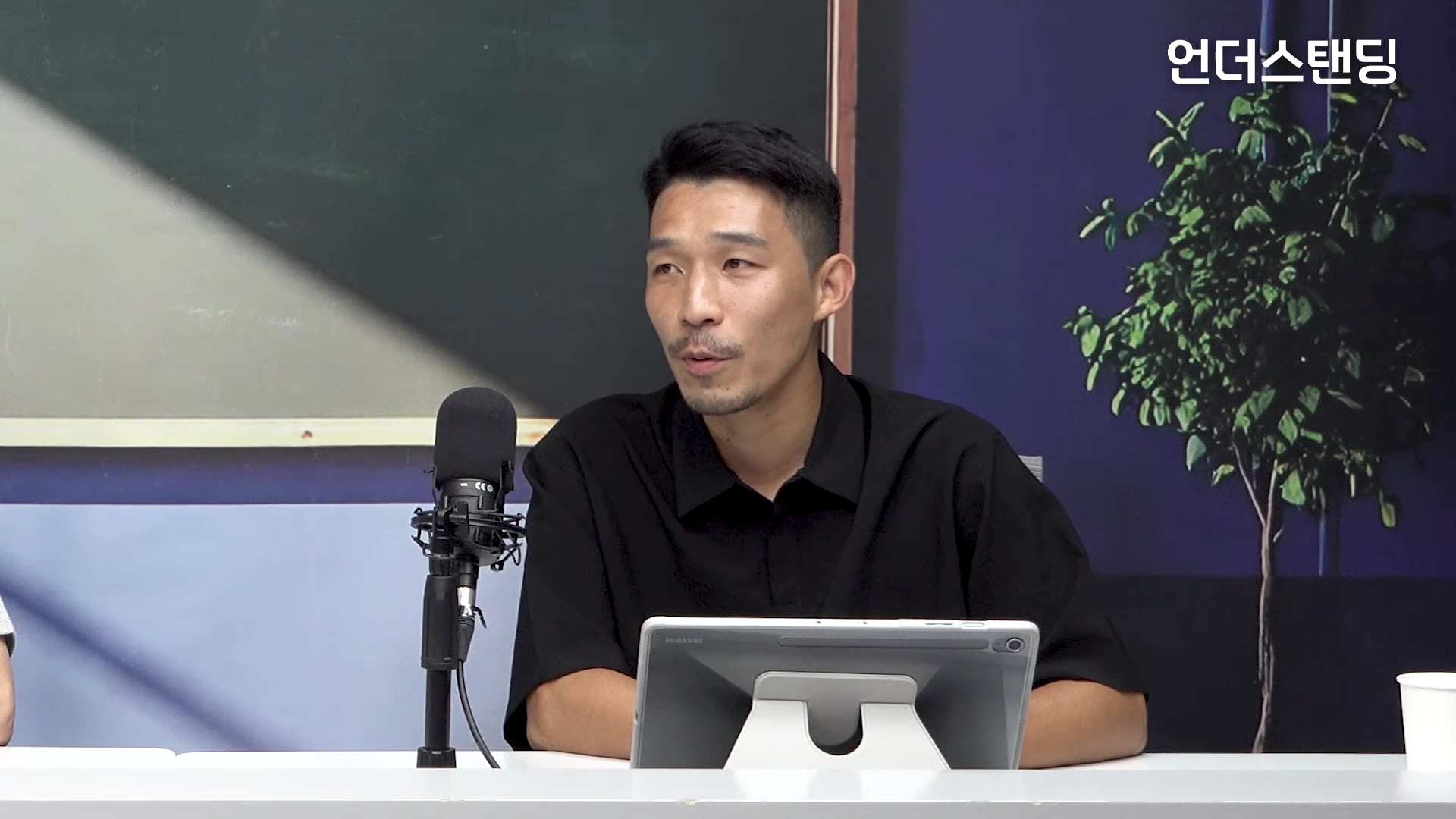 스튜디오 책상 앞에 앉아 검은색 반소매 셔츠를 입은 남성이 마이크를 앞에 두고 이야기를 나누고 있는 모습이다.