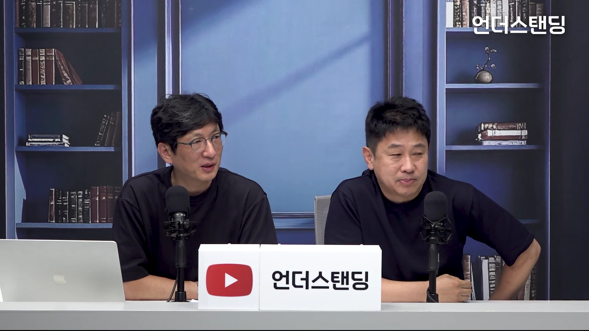 서재 배경 앞에서 검은색 상의를 입은 두 명의 남성이 책상 앞에 앉아 각각 마이크를 앞에 두고 대화를 나누고 있는 스튜디오 장면입니다.