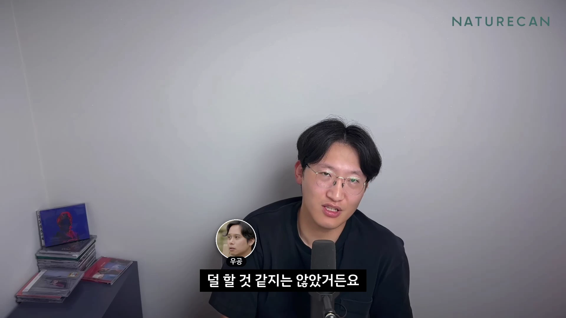 안경을 쓴 남성이 마이크 앞에 앉아 정면을 바라보며 이야기하고 있으며, 화면 하단에는 그의 이름이 적힌 작은 원형 프로필 사진과 자막이 떠 있습니다.