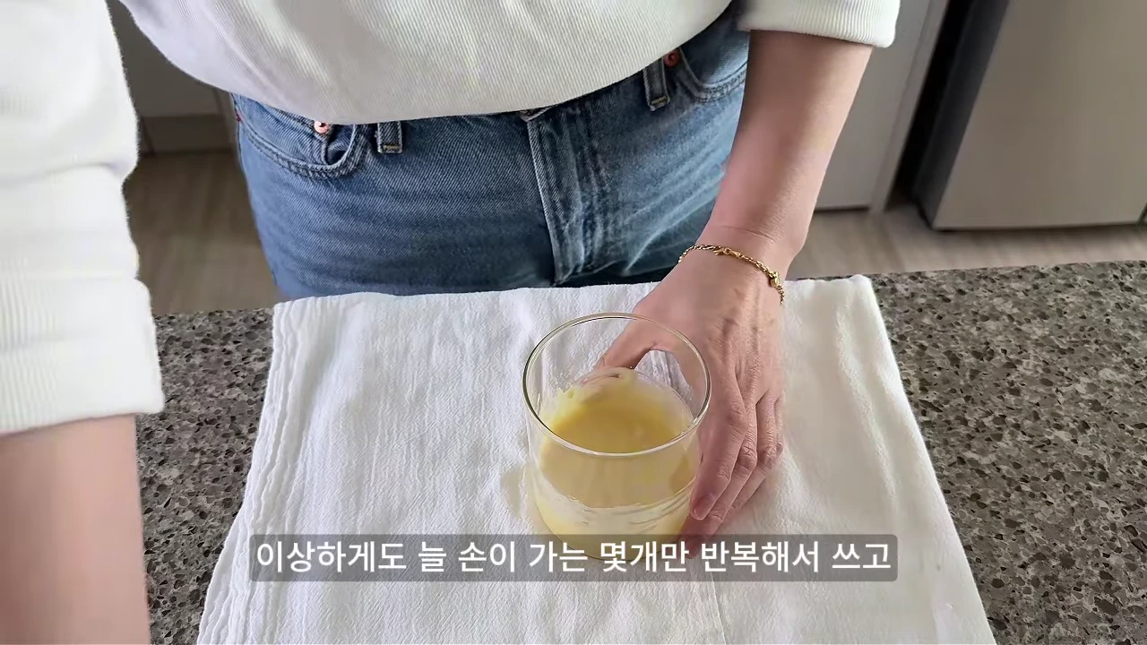 흰색 상의와 청바지를 입은 사람이 주방 상판 위 흰 천 위에 놓인 유리컵을 잡고 있는 모습이 담긴 영상 캡처 화면입니다.