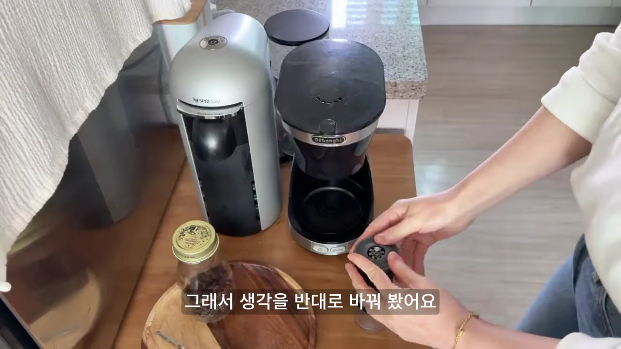 주방 조리대 위에 두 대의 커피 머신과 원두 병이 놓여 있고, 한 사람이 손으로 소형 커피 도구를 조작하고 있는 모습