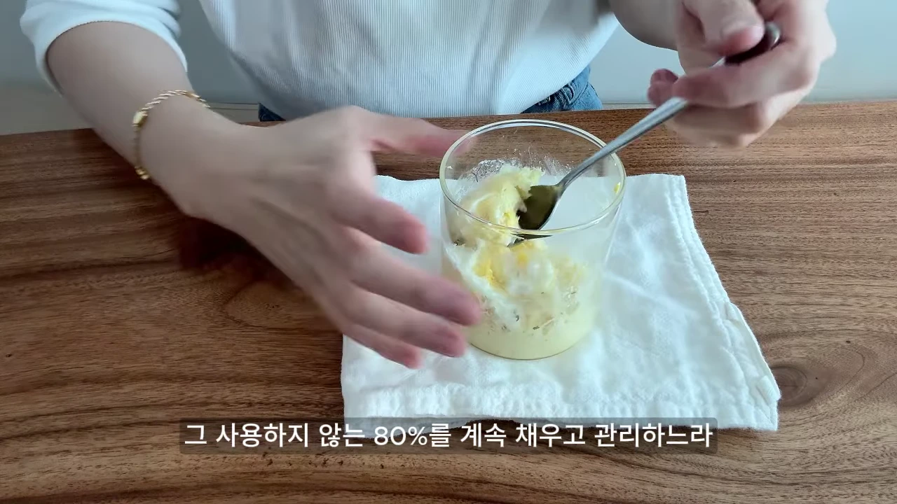 나무 식탁 위에 흰 천을 깔고 그 위에 놓인 투명한 유리컵에 담긴 음식을 숟가락으로 저으며 먹는 사람의 손과 상반신.