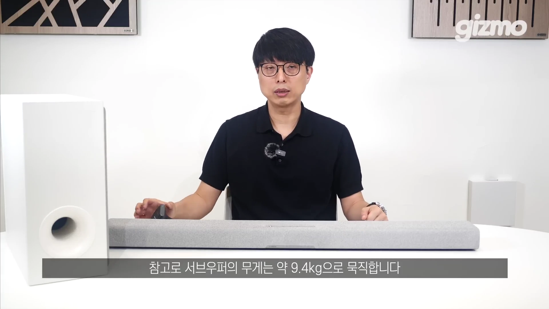 흰색 배경의 스튜디오에서 안경을 쓴 남성이 야마하 사운드바 본체 옆에 서브우퍼를 두고 설명하는 모습