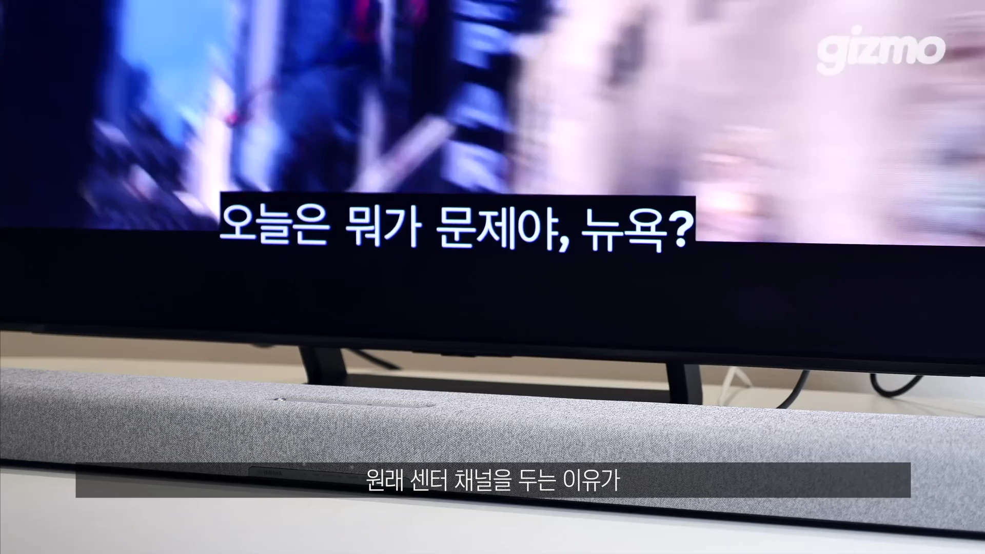 회색 패브릭 소재의 긴 사운드바가 TV 바로 아래에 놓여 있으며, 화면에는 자막이 떠 있습니다.