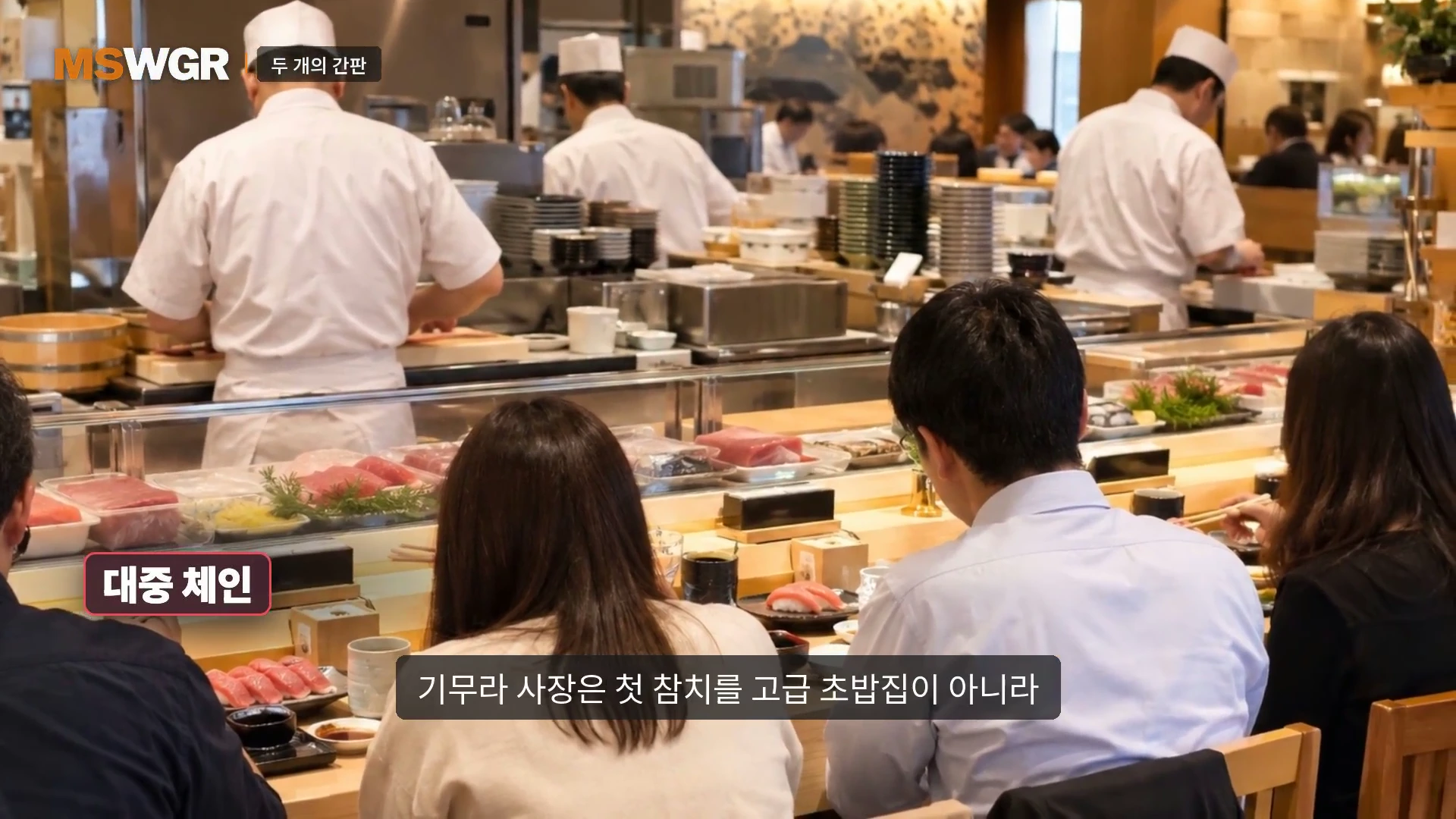 흰색 조리복을 입은 요리사들이 뒤에서 초밥을 만들고, 앞쪽 바 테이블에 앉은 손님들이 초밥을 즐기고 있는 대중적인 초밥 전문점의 모습.