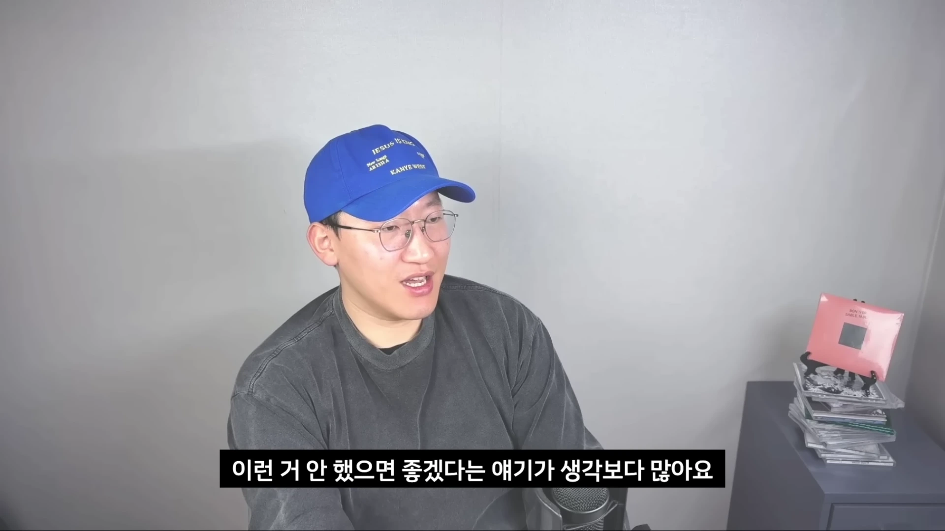 파란색 야구 모자를 쓰고 안경을 쓴 남성이 밝은 회색 벽을 배경으로 앉아 이야기하고 있으며, 화면 하단에는 한국어 자막이 표시되어 있다.