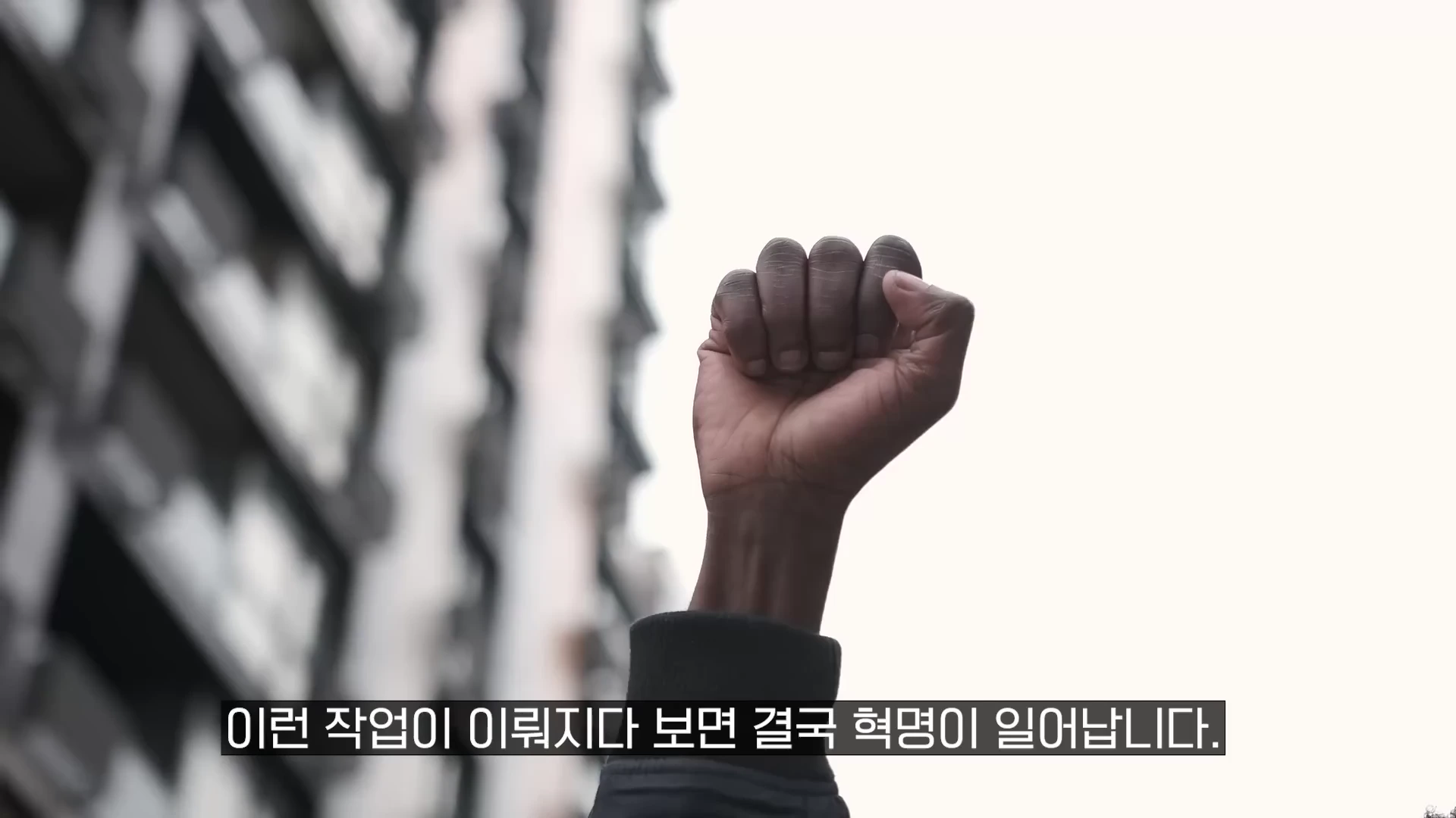하늘을 향해 높이 쳐든 사람의 주먹과 그 아래로 보이는 흐릿한 건물 외벽, 화면 하단에는 흰색 자막이 배치되어 있습니다.