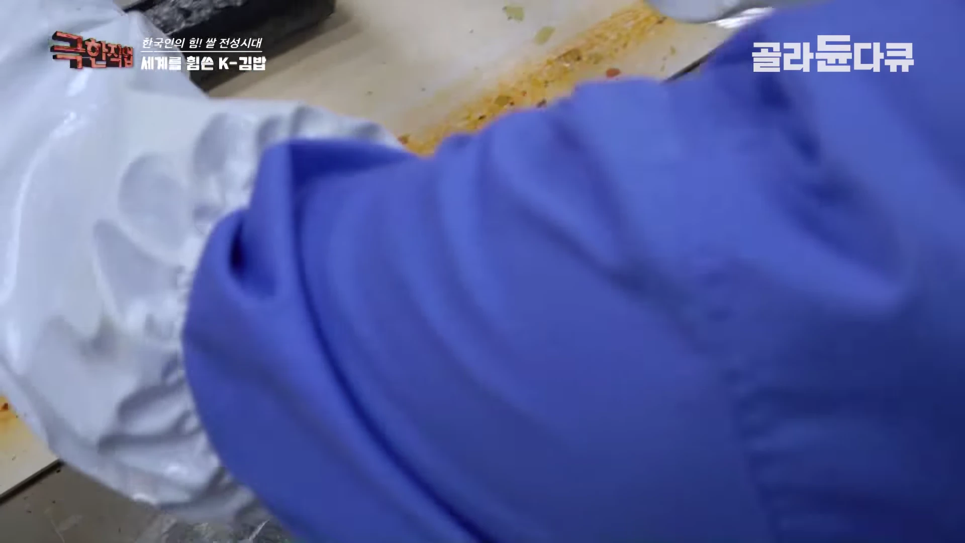 작업자가 위생 장갑을 끼고 김밥의 재료를 조심스럽게 말아 올리는 모습이 담긴 화면이다.