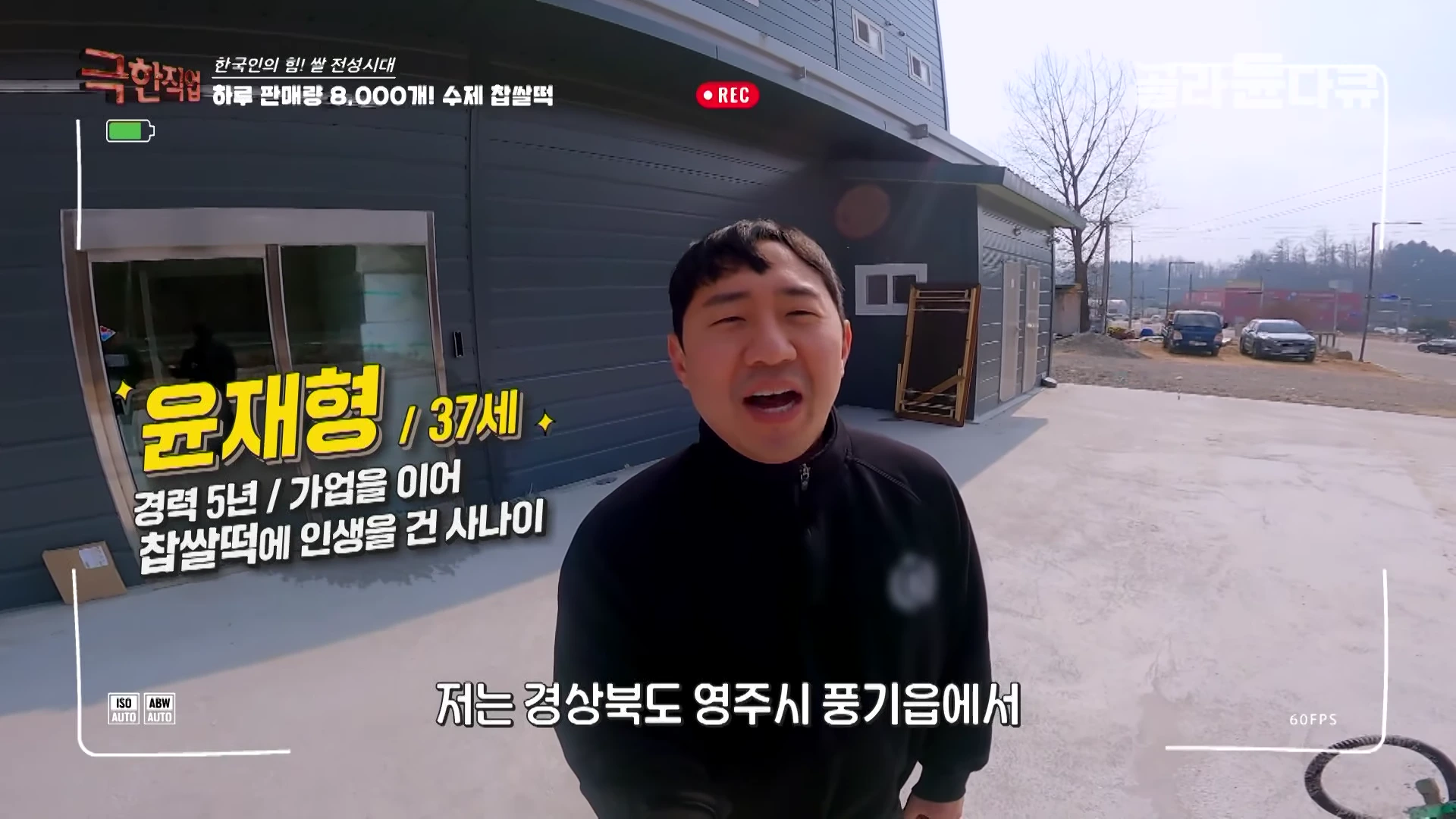 실외에서 짙은 회색 건물 앞에 서서 카메라를 바라보며 이야기하고 있는 37세 남성