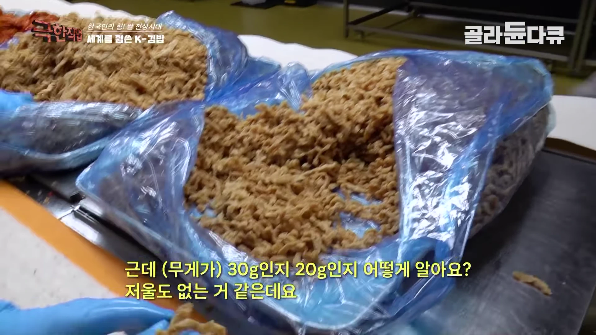 비닐에 담긴 조림 유부와 당근 등 김밥 재료가 트레이에 놓여 있고, 파란색 위생 장갑을 낀 작업자의 손이 재료를 집는 모습. 화면 하단에는 무게를 재는 저울이 없다는 내용의 노란색 자막이 표시되어 있다.