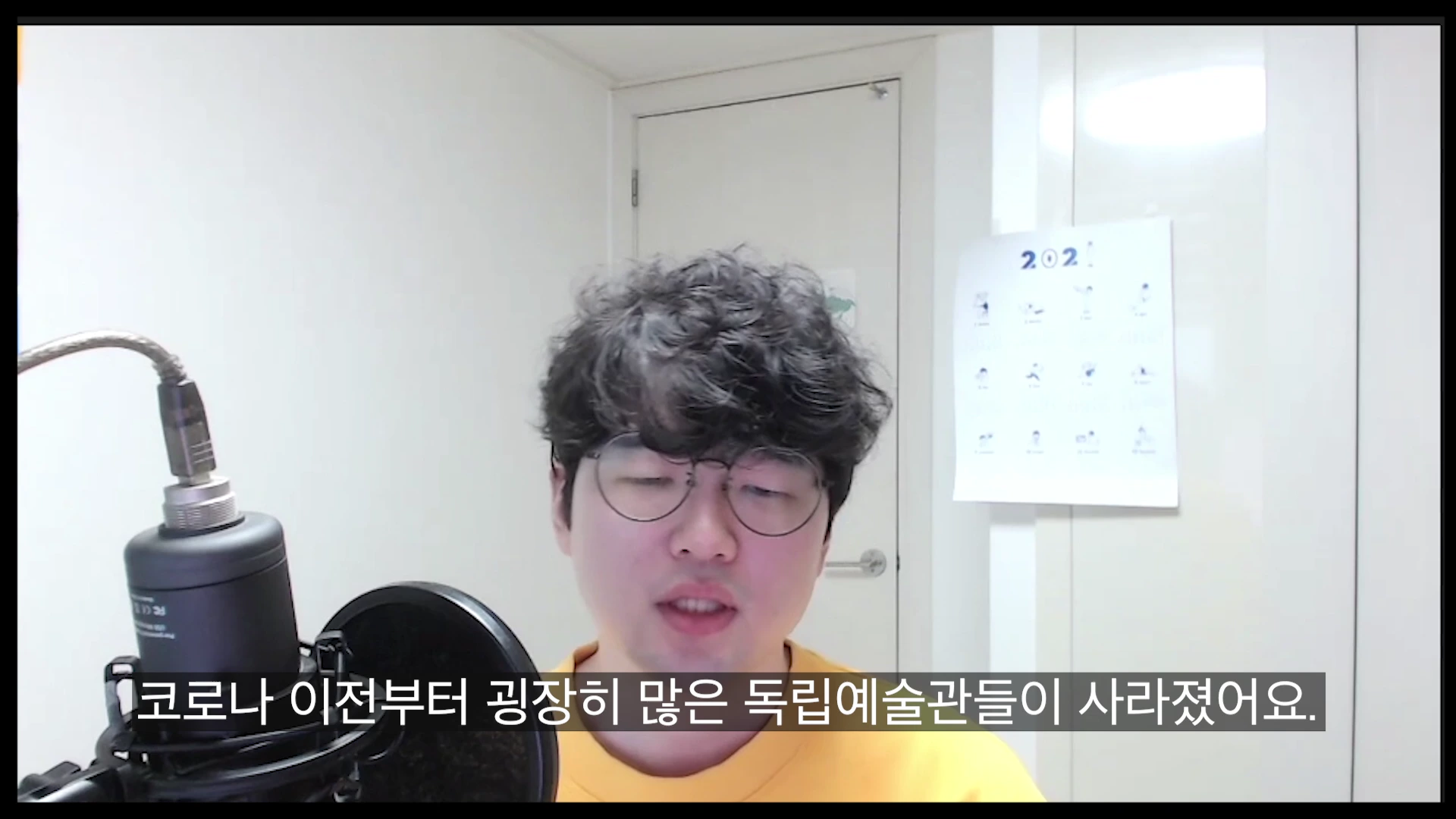 흰색 배경의 방 안에서 노란색 티셔츠를 입고 안경을 쓴 남성이 마이크 앞에 앉아 대화하고 있는 모습. 화면 하단에는 '코로나 이전부터 굉장히 많은 독립예술관들이 사라졌어요.'라는 한글 자막이 표시되어 있다.