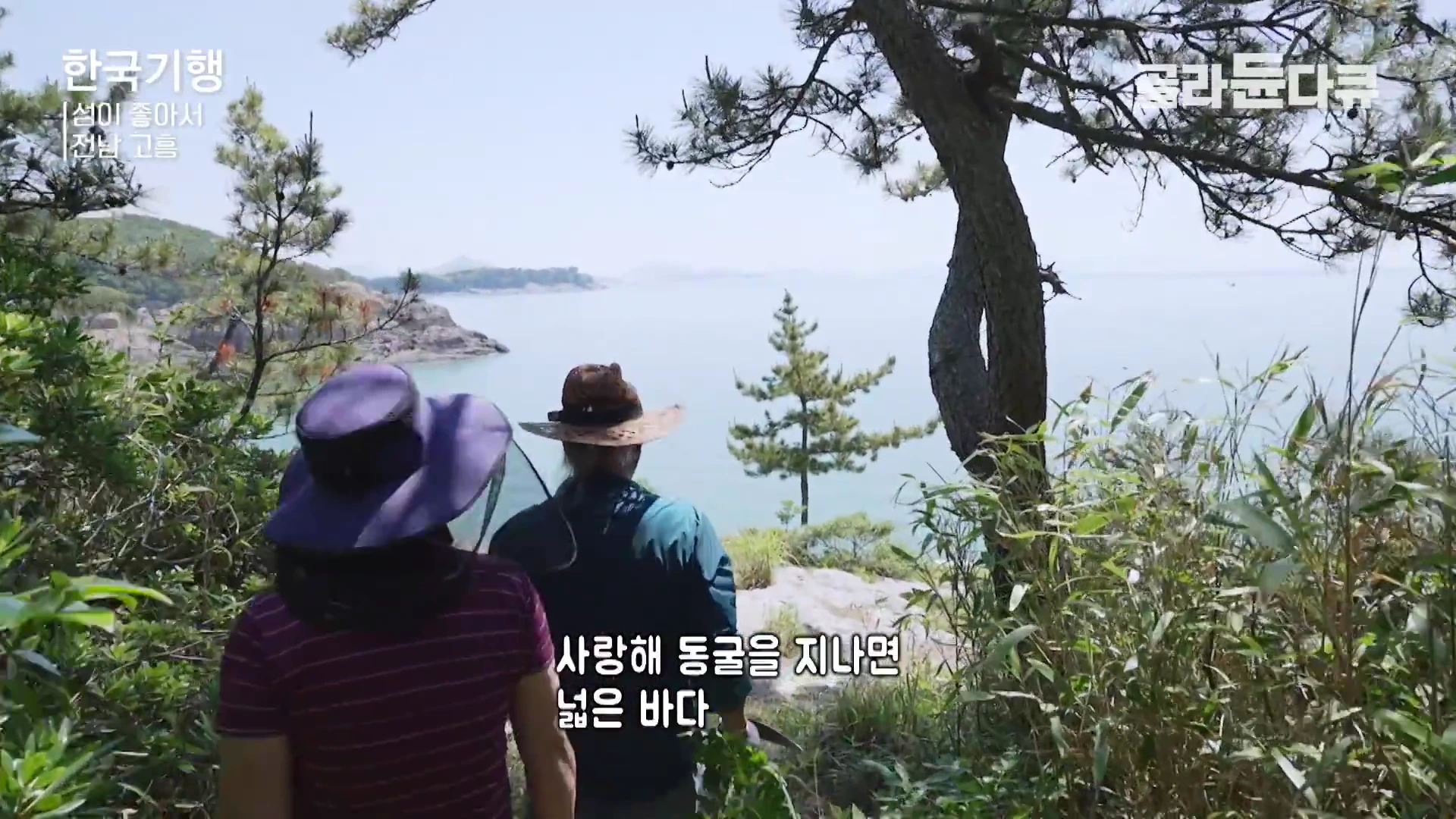 모자를 쓴 중년 부부가 나무가 우거진 오솔길을 따라 걸으며 앞에 펼쳐진 잔잔한 바다를 바라보고 있는 뒷모습입니다.