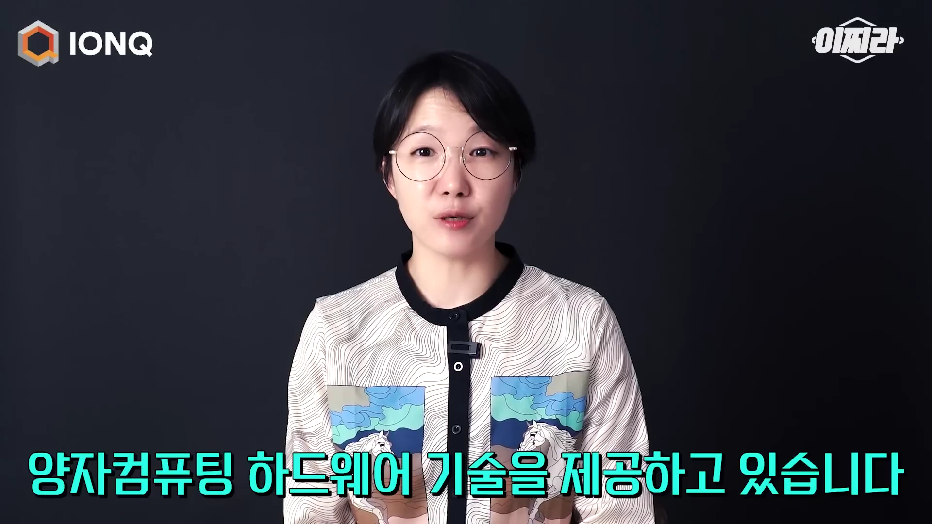 안경을 쓴 여성 진행자가 검은색 배경 앞에서 정면을 바라보며 이야기하고 있고, 화면 하단에는 '양자컴퓨팅 하드웨어 기술을 제공하고 있습니다'라는 자막이 표시되어 있습니다.
