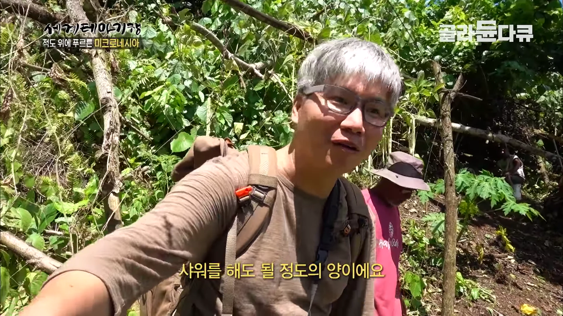 안경을 쓴 중년 남성이 울창한 숲길을 걸으며 카메라를 보고 이야기하고 있고, 그 뒤로 모자를 쓴 다른 남성이 함께 걷고 있는 모습