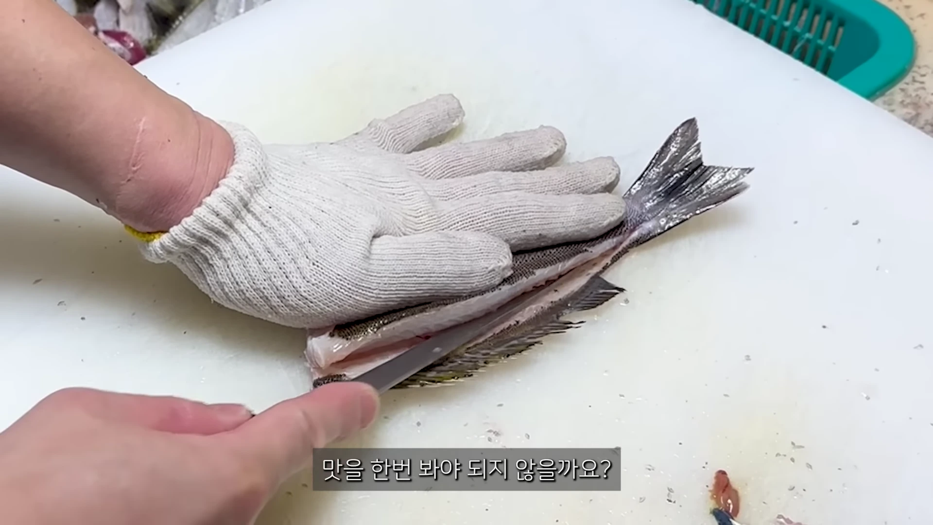 흰색 도마 위에서 작업용 장갑을 낀 손이 생선의 몸통을 누르고 칼로 살을 발라내는 모습
