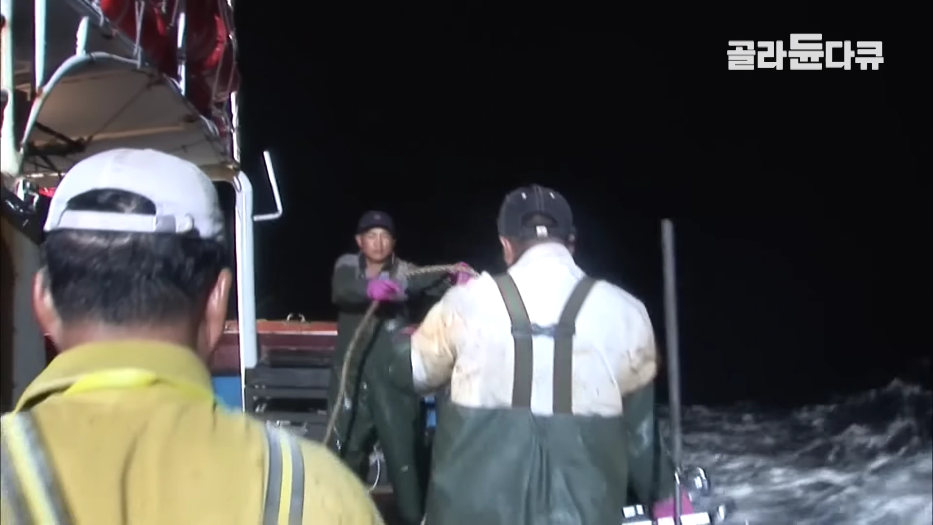 어두운 밤, 바다 위 선박에서 모자와 작업복을 착용한 선원들이 굵은 밧줄을 잡아당기며 조업에 집중하고 있는 모습.