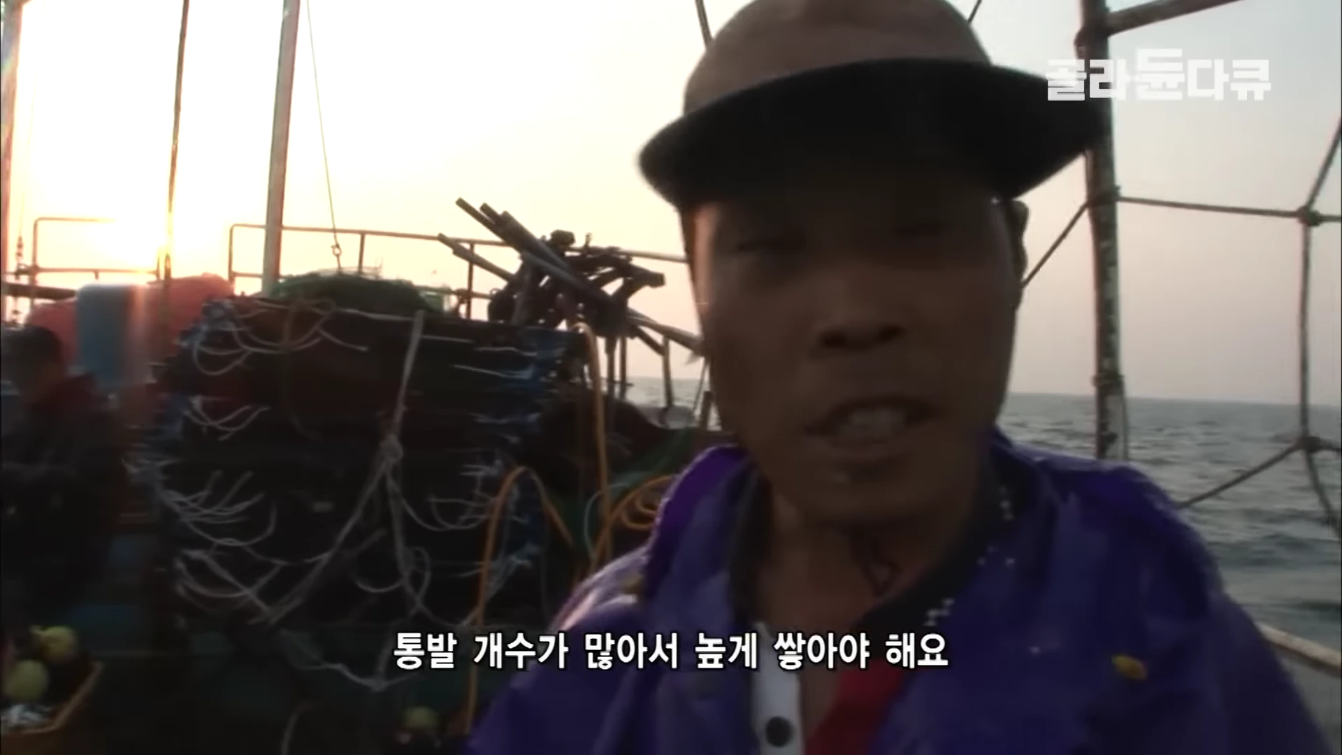 모자를 쓴 어부가 갑판 위에서 바다를 배경으로 인터뷰하고 있으며, 뒤쪽에는 높게 쌓인 통발이 보임