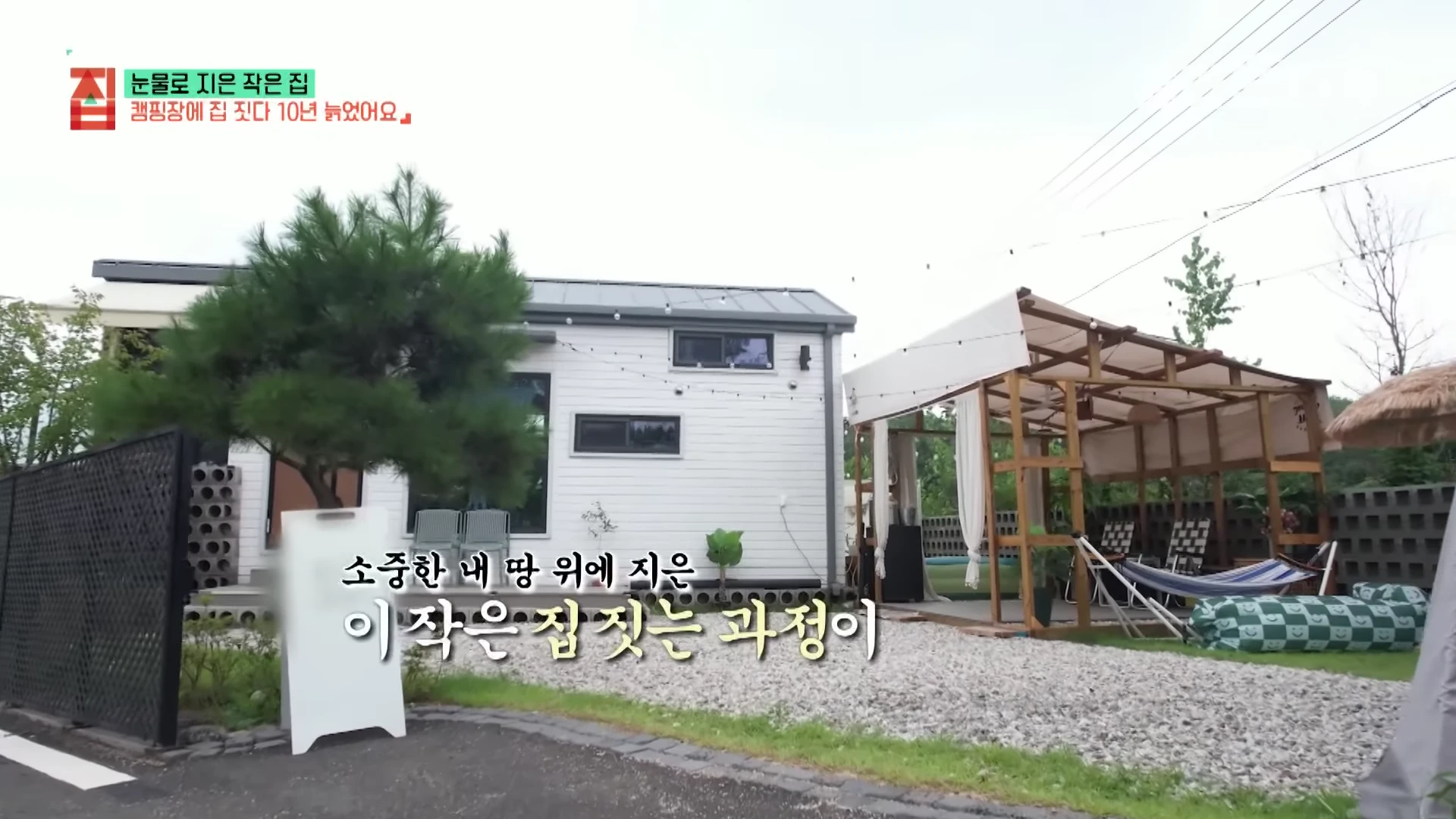 캠핑장 부지 위에 자리 잡은 하얀색 박공지붕의 소형 이동식 주택과 옆에 설치된 나무 골조의 야외 파고라 및 해먹이 보입니다.