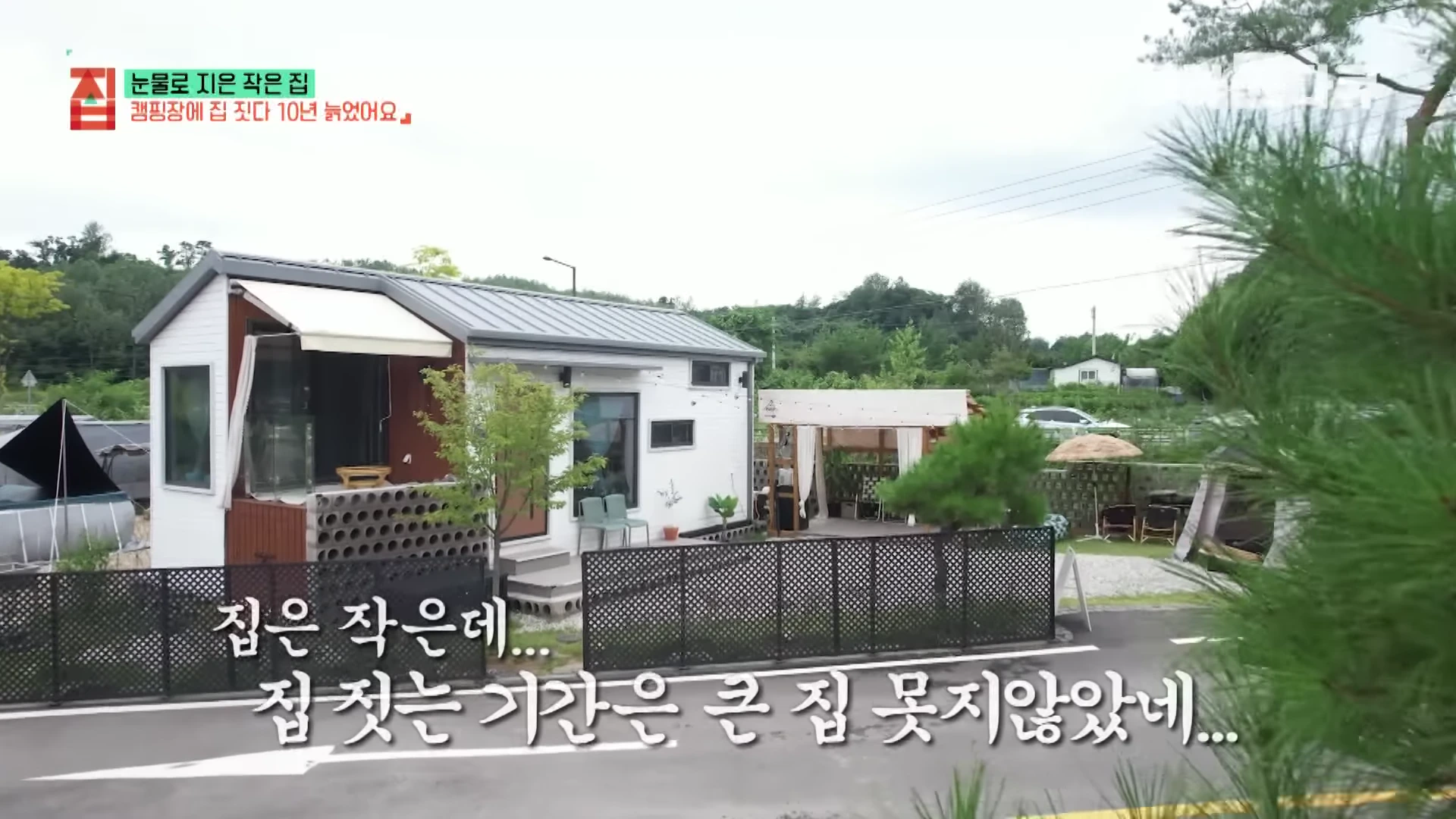 캠핑장 내에 자리 잡은 박공지붕 형태의 소형 이동식 주택과 주택 앞 울타리, 주변 풍경이 보이는 영상 캡처 화면.