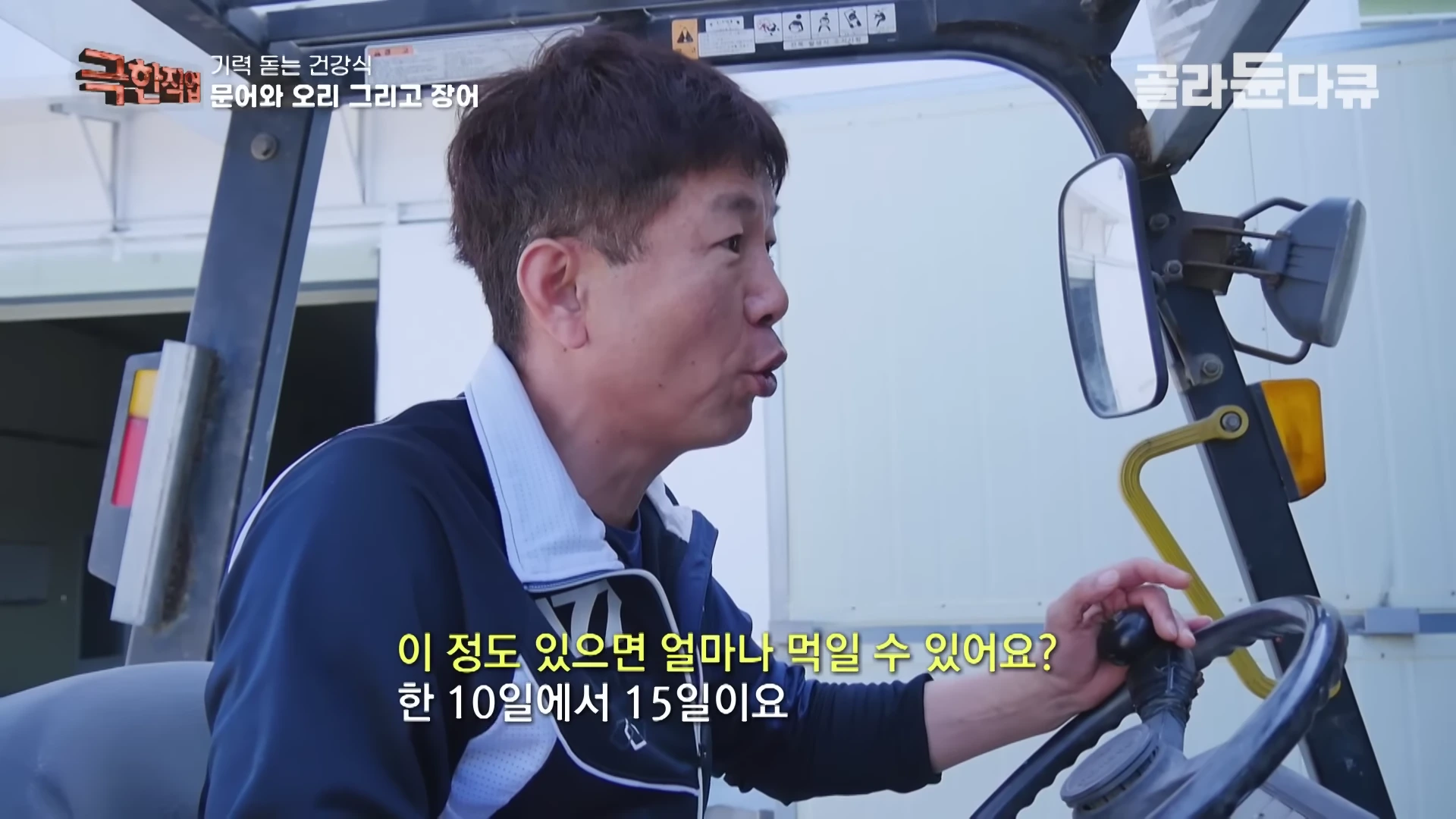 지게차 운전석에 앉아 있는 중년 남성이 한 손으로 운전대를 잡고 측면을 바라보며 인터뷰에 응하고 있는 모습이다.