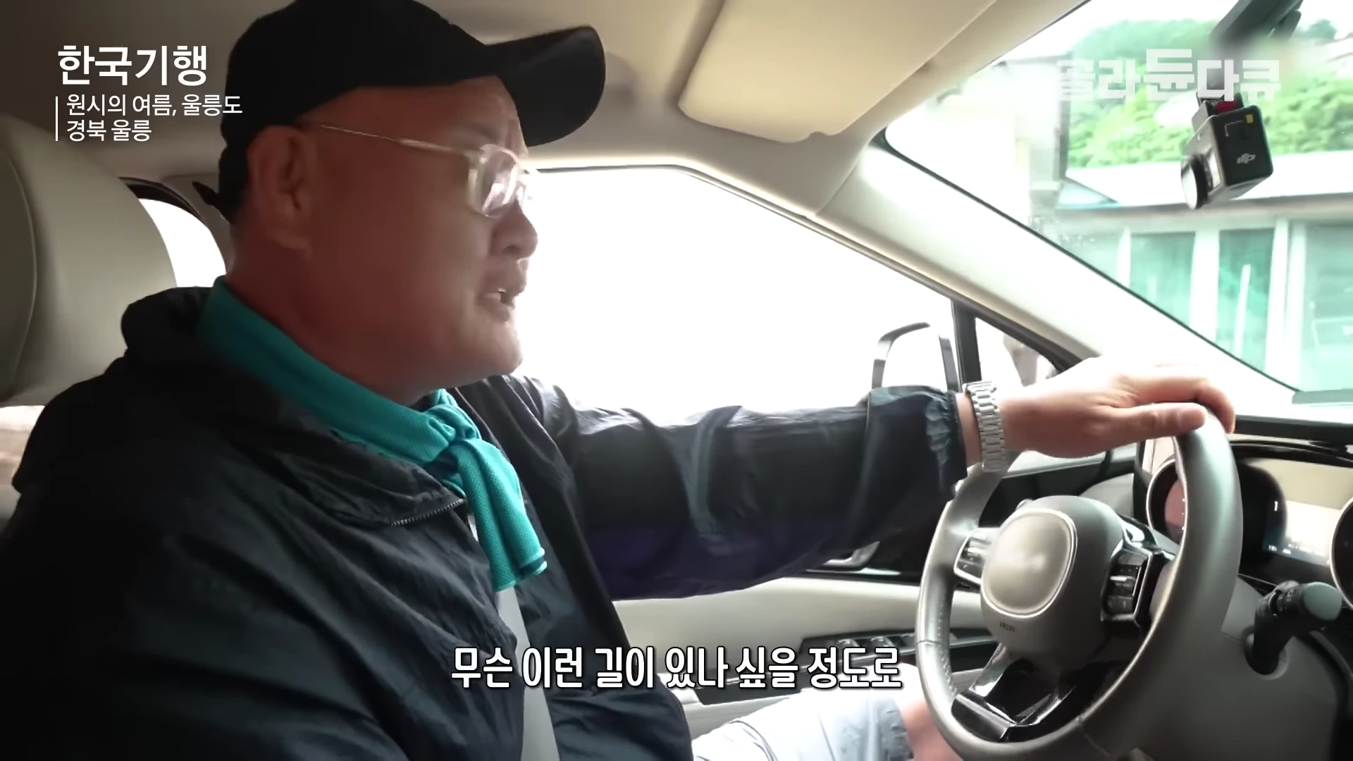 안경과 모자를 쓴 남성이 차량 운전석에서 운전대를 잡고 앞을 보며 이야기하고 있는 모습