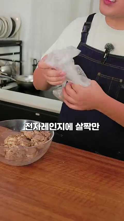 앞치마를 입은 요리사가 투명한 비닐봉지를 열어 투명한 유리 볼에 담긴 고기를 꺼내고 있으며 화면 중앙에는 전자레인지에 살짝만이라는 자막이 떠 있습니다.