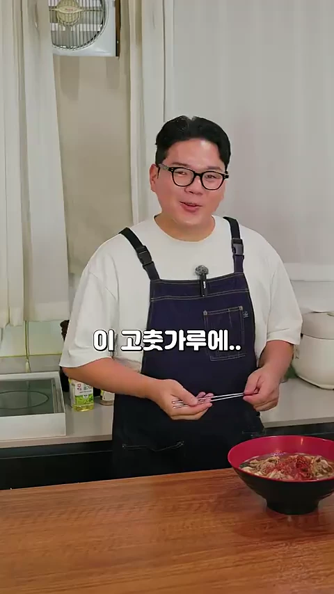 흰색 반소매 티셔츠에 남색 앞치마를 두른 안경 쓴 남성이 주방 조리대 앞에서 젓가락을 들고 이야기하고 있으며, 앞에는 완성된 요리가 담긴 그릇이 놓여 있다.