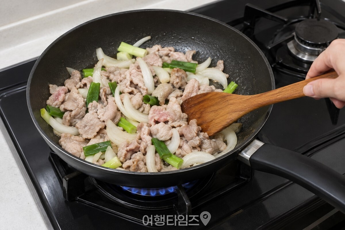 검은 팬에서 겉면이 익기 시작한 돼지고기에 양파와 대파를 넣어 함께 볶고 있다. / 기사 이해를 돕기 위한 생성형 이미지
