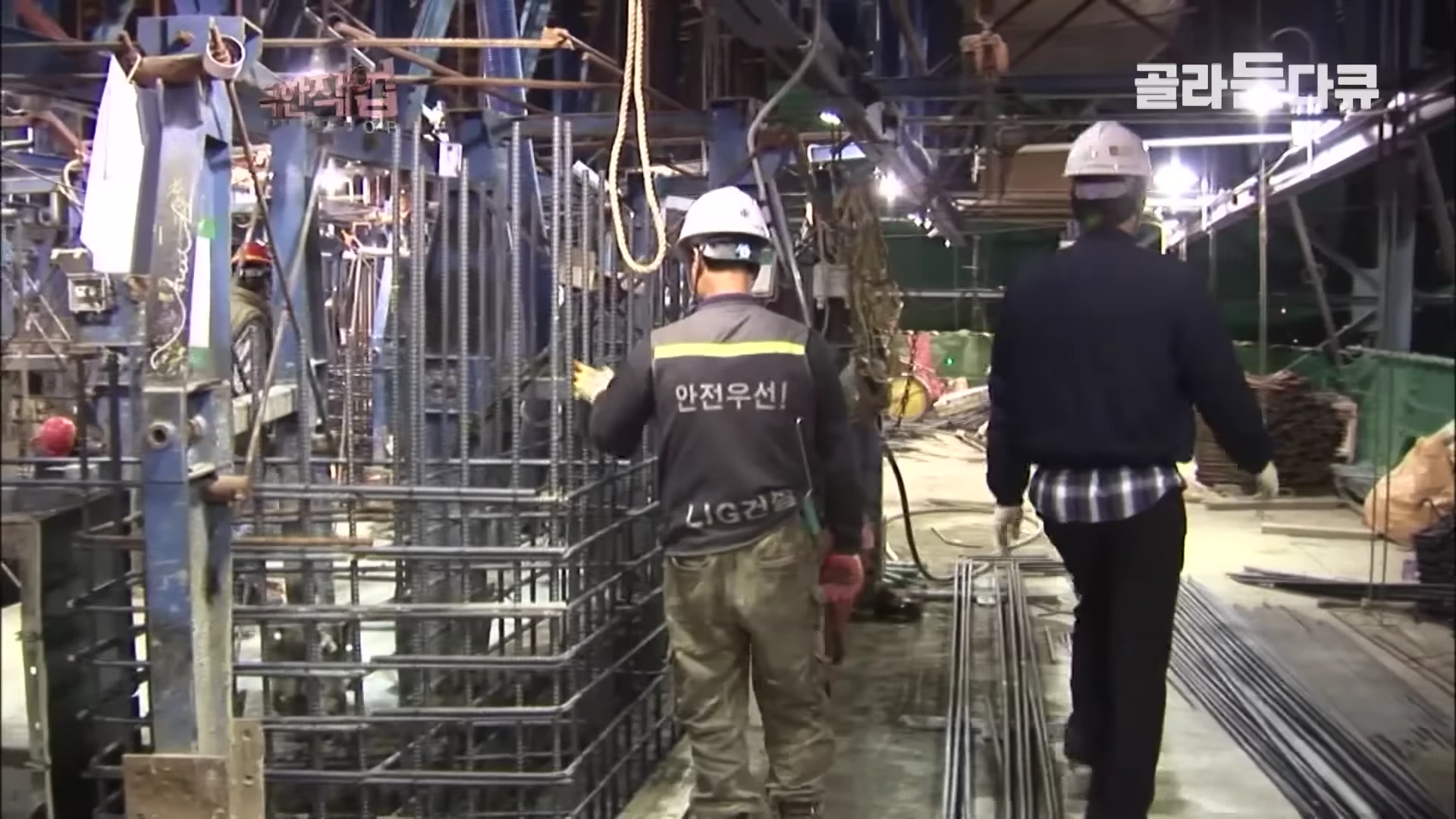 건설 현장에서 안전모를 쓴 두 명의 작업자가 철근 구조물 사이를 지나가며 작업 상태를 확인하고 있다.
