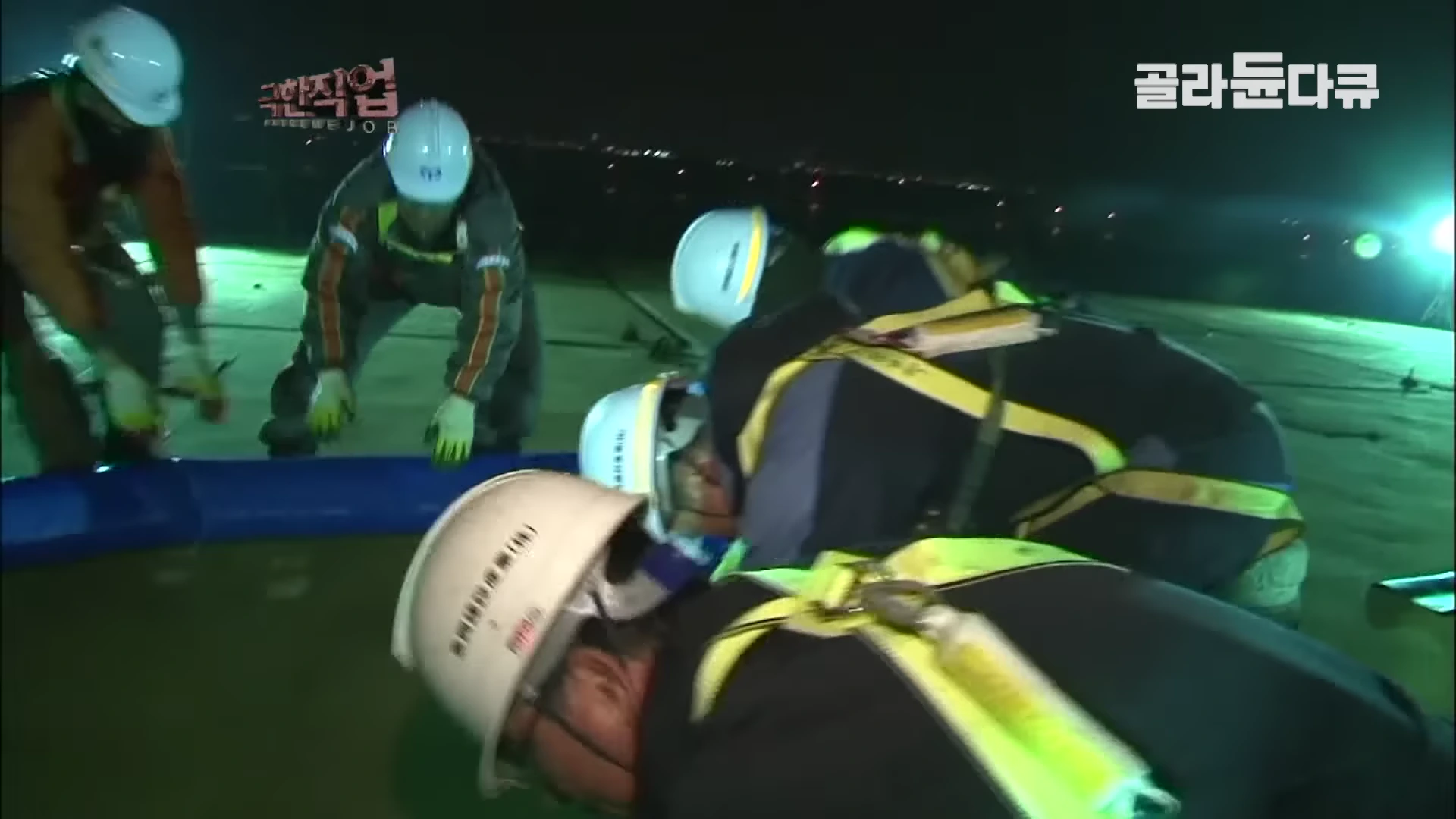 흰색 안전모와 형광색 안전 조끼를 착용한 여러 명의 작업자가 야간에 어두운 바닥 위에서 무릎을 굽히고 앉아 장비를 점검하고 있다.