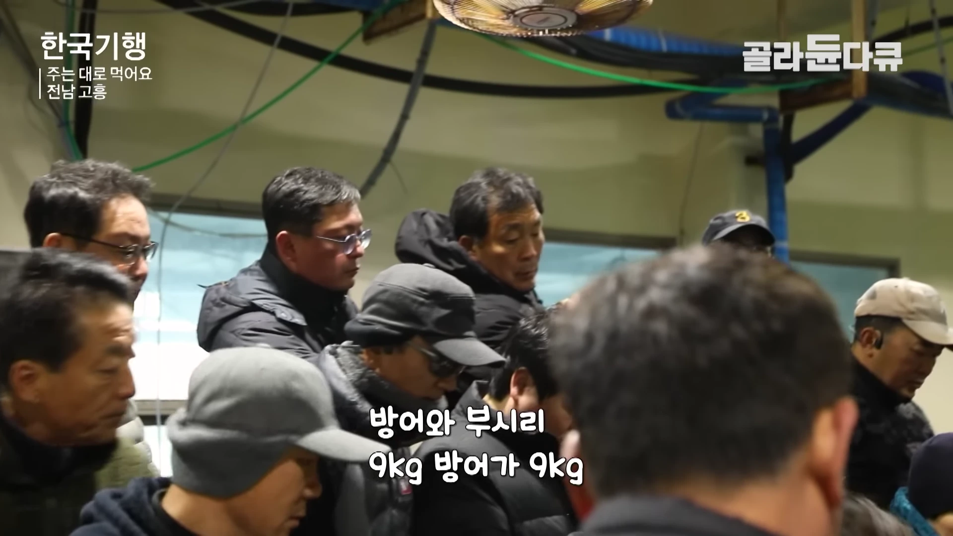 실내 위판장에서 모자나 안경을 쓴 여러 명의 남성들이 방어와 부시리 경매 과정을 지켜보고 있다.