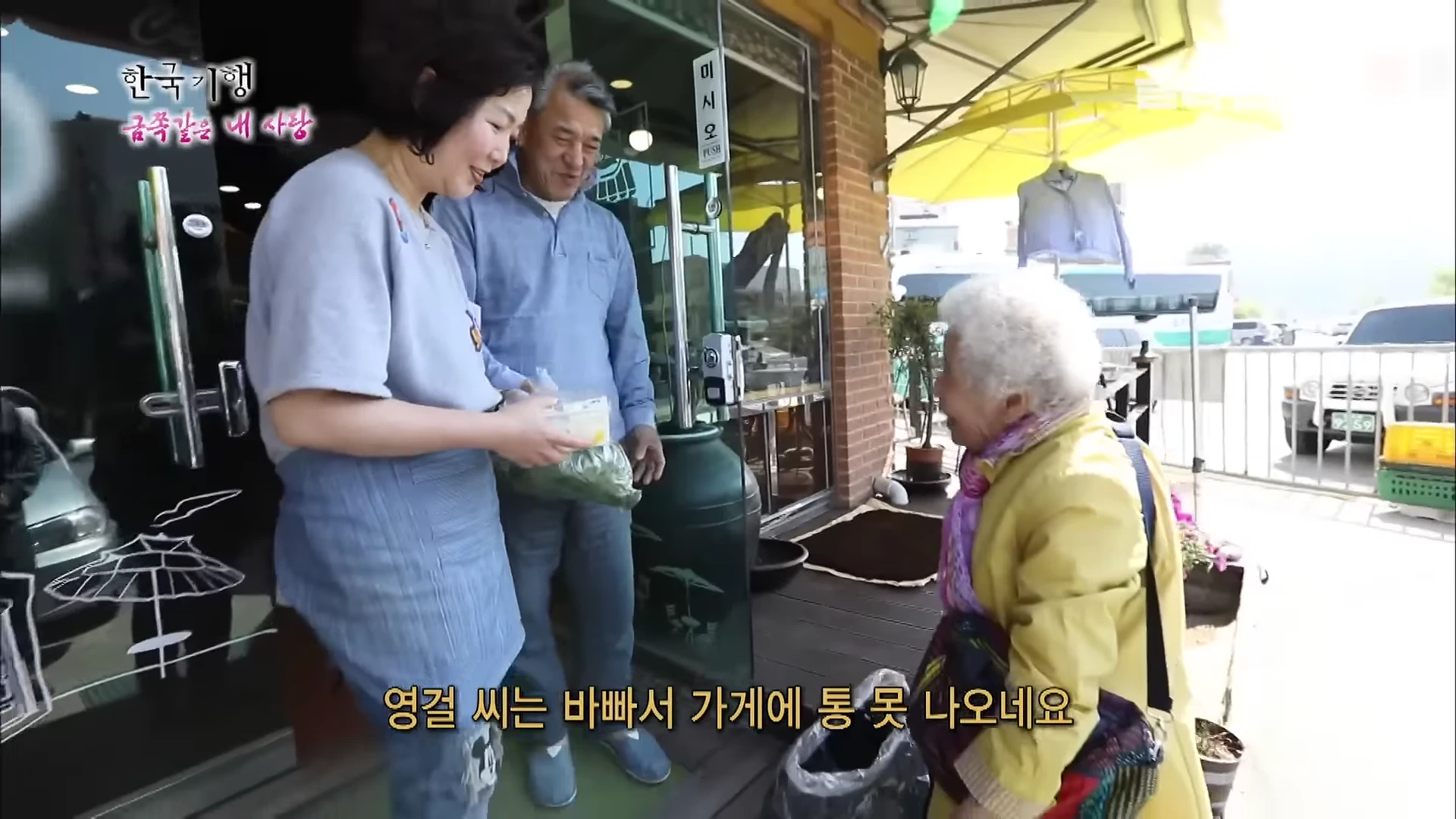 가게 앞에서 밝은 노란색 재킷을 입은 백발의 할머니가 앞치마를 두른 여성과 서서 이야기를 나누고 있으며, 그 뒤로 편안한 옷차림의 남성이 미소 짓고 있다.
