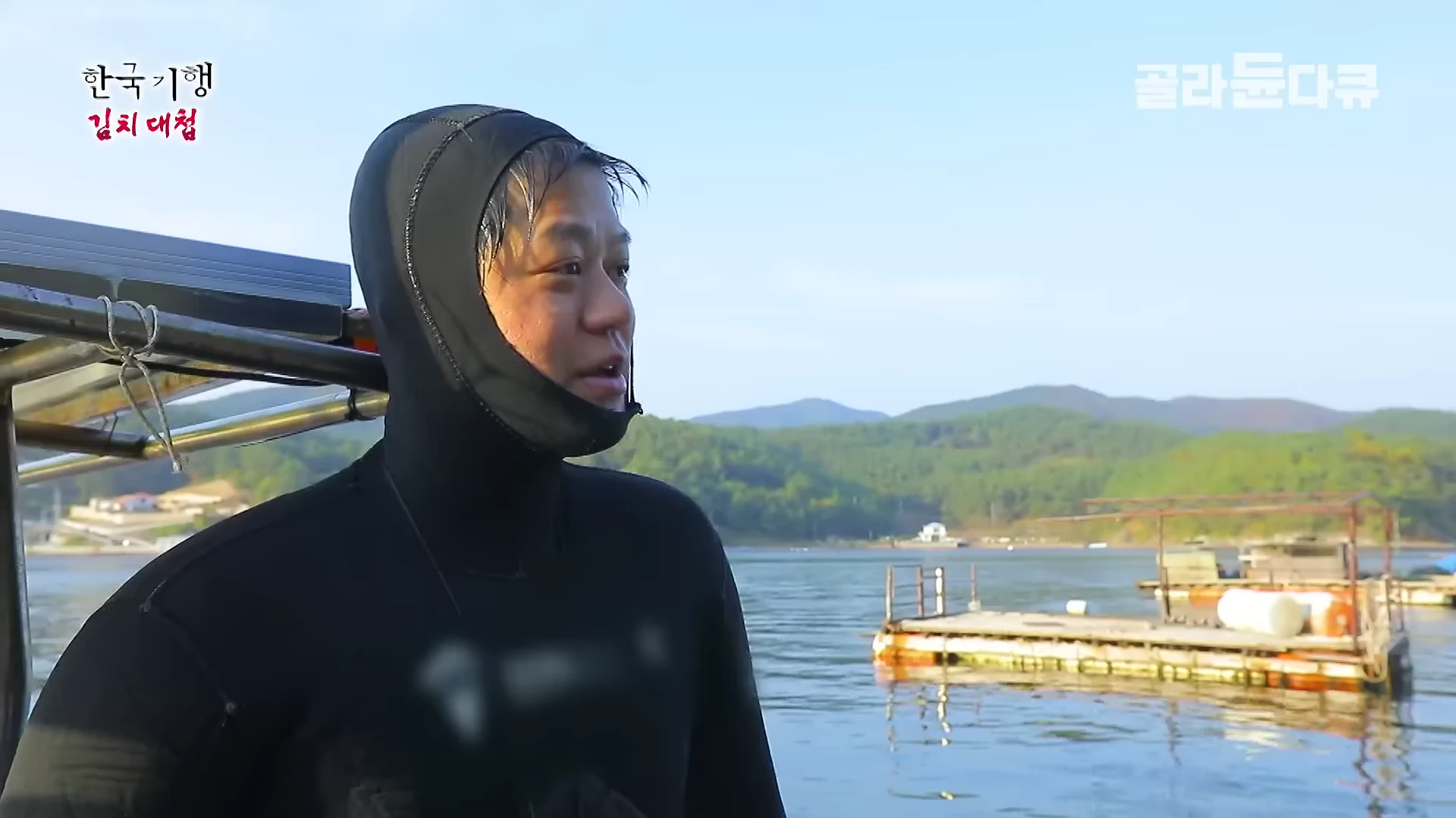 잠수복 후드를 쓴 남성이 배 위에서 바다를 바라보고 있으며 뒤편으로 잔잔한 바다와 산이 펼쳐진 모습