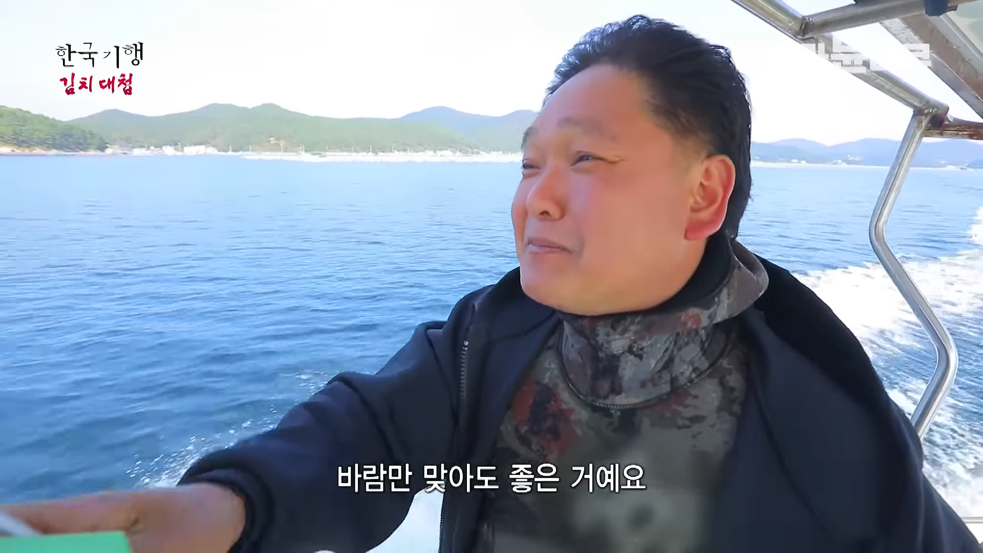 잠수복을 입은 남성이 달리는 배 위에서 바다 바람을 맞으며 환하게 웃고 있는 모습