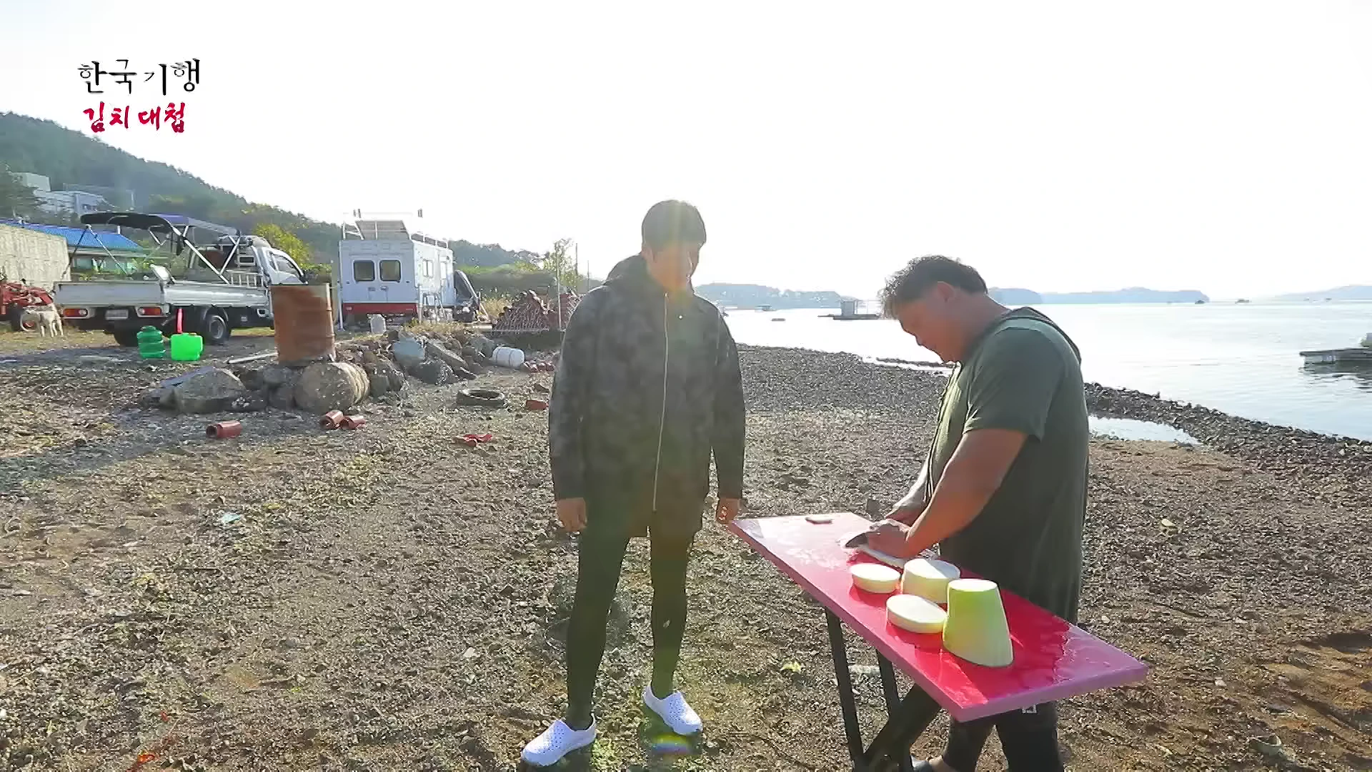 바닷가 공터에서 한 남성이 야외 테이블에 놓고 무를 썰고 있고, 옆에 다른 남성이 서서 이를 지켜보는 장면.