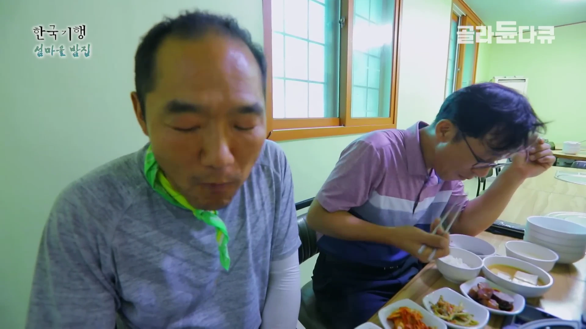 실내 식당에서 두 남성이 식탁에 차려진 여러 가지 반찬과 밥을 먹고 있는 모습입니다.