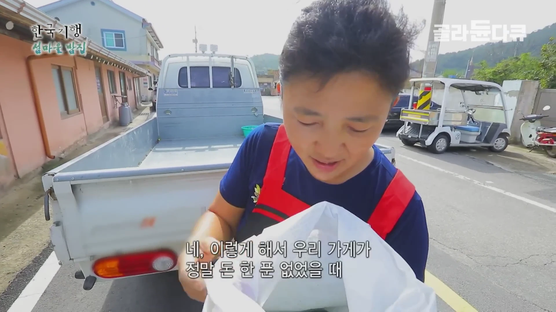 붉은색 앞치마를 두른 여성이 흰색 비닐봉지를 들고 이야기하고 있으며, 뒤쪽에는 소형 트럭과 골프 카트가 주차되어 있다.