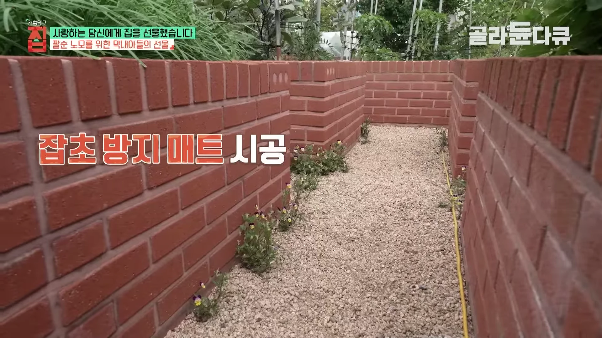 붉은 벽돌로 쌓은 낮은 담장 사이로 자갈이 깔린 통로가 보이며, '잡초 방지 매트 시공'이라는 한글 자막이 화면 중앙에 나타나 있습니다.
