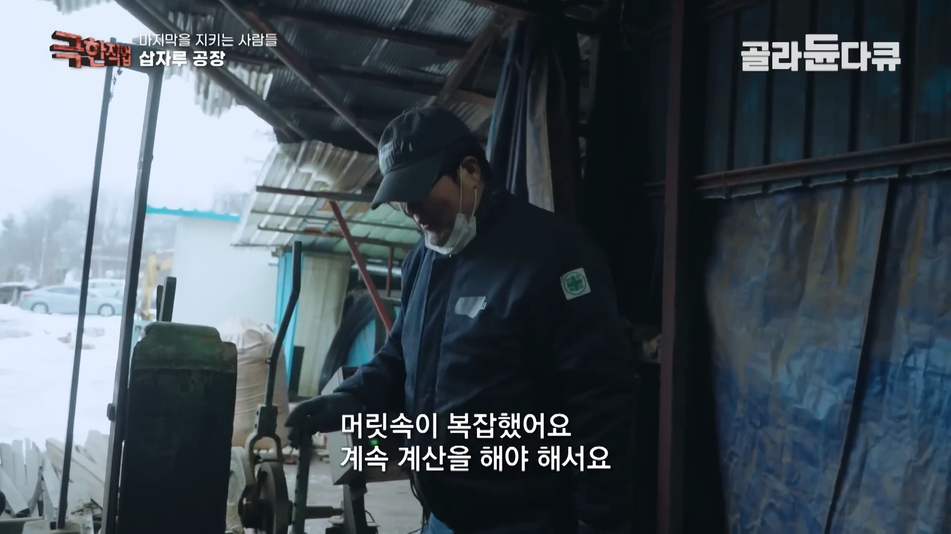작업장에서 모자를 쓴 남성이 삽자루 제작 기계 앞에서 무언가를 확인하며 작업하고 있는 모습