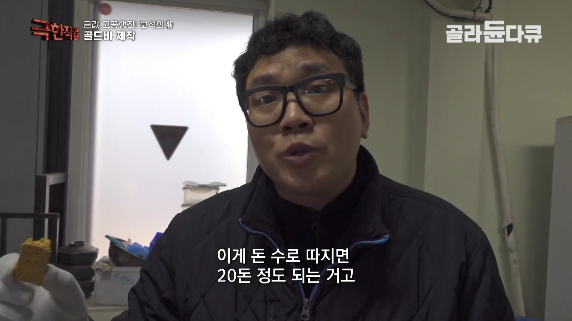 안경을 쓴 남성이 흰 장갑을 낀 손으로 작은 금덩어리를 들고 설명하는 모습이 담긴 영상 캡처 화면입니다.