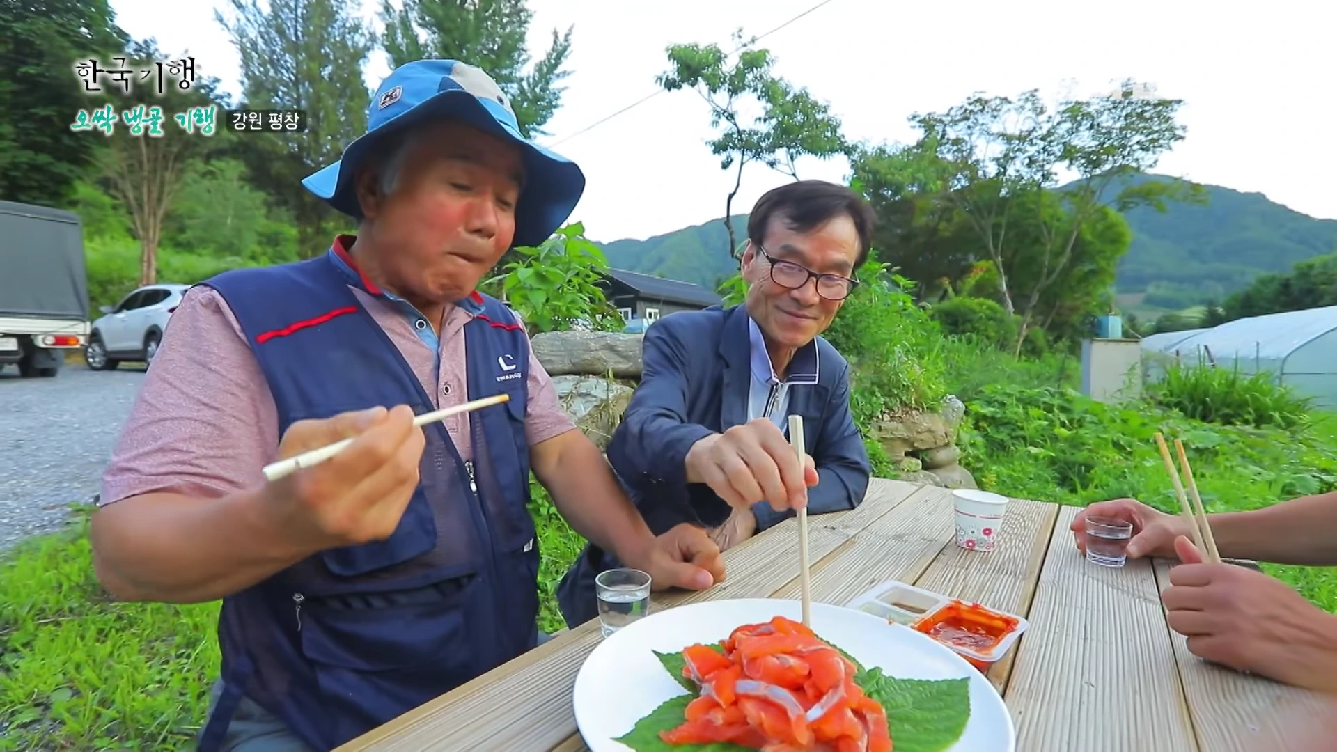 두 남자가 야외 테이블에 둘러앉아 깻잎 위에 올려진 붉은 송어회를 젓가락으로 집어 먹는 모습.