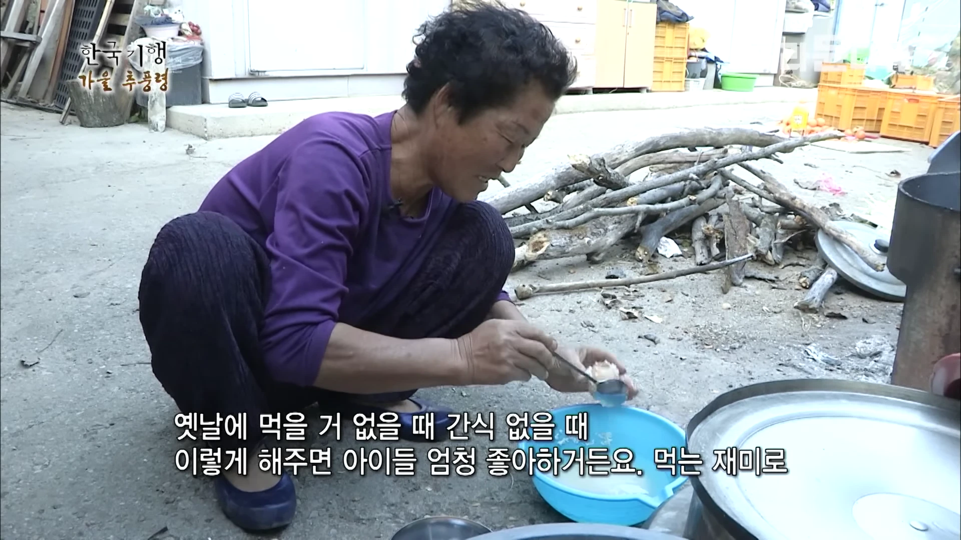 마당에서 보라색 상의를 입은 노인이 쪼그리고 앉아 파란색 바구니에 담긴 달걀을 숟가락으로 만지고 있으며, 주변에는 땔감이 쌓여 있다.