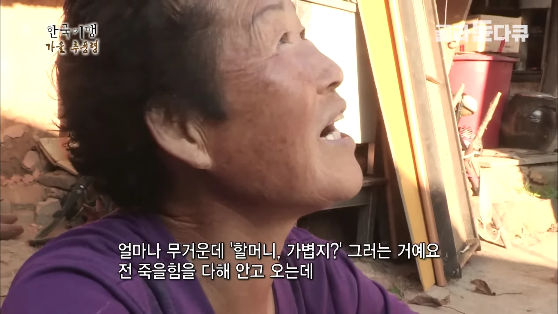 보라색 상의를 입은 노년의 여성이 하늘을 바라보며 환하게 웃고 있는 얼굴 클로즈업 장면이며, 화면 하단에는 자막이 포함되어 있습니다.