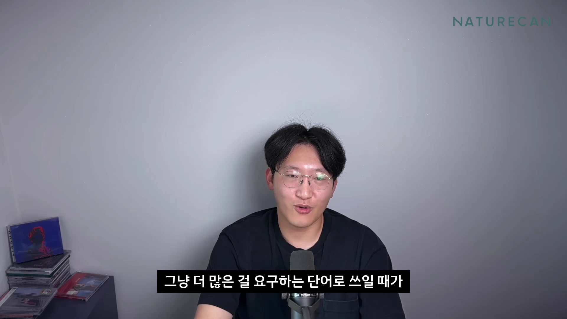 검은색 티셔츠를 입고 안경을 쓴 남성이 마이크 앞에서 이야기하고 있으며, 화면 하단에는 '그냥 더 많은 걸 요구하는 단어로 쓰일 때가'라는 자막이 표시된 영상 캡처 화면입니다.