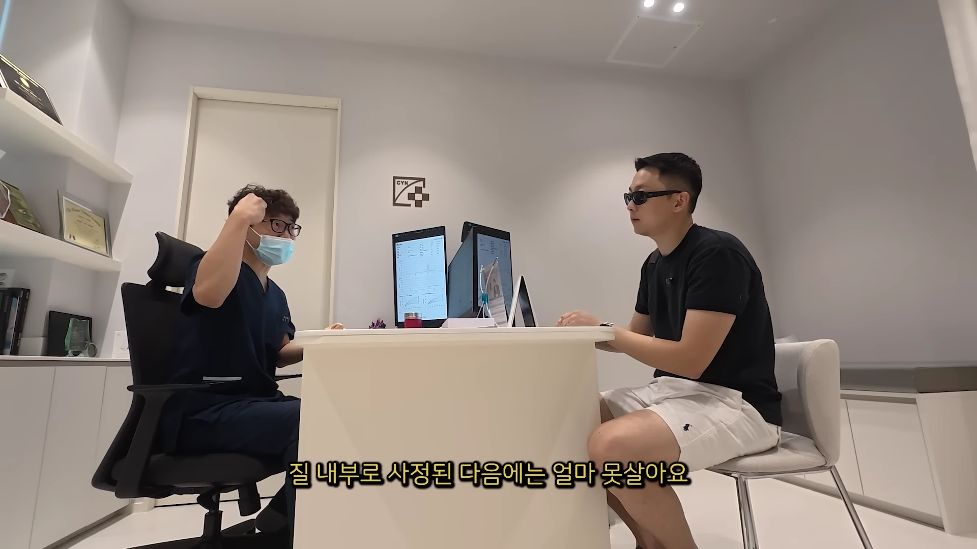 병원 진료실에서 파란색 수술복을 입은 의사와 검은색 티셔츠를 입고 선글라스를 쓴 남성이 마주 앉아 상담하고 있으며, 화면 하단에는 자막이 있습니다.