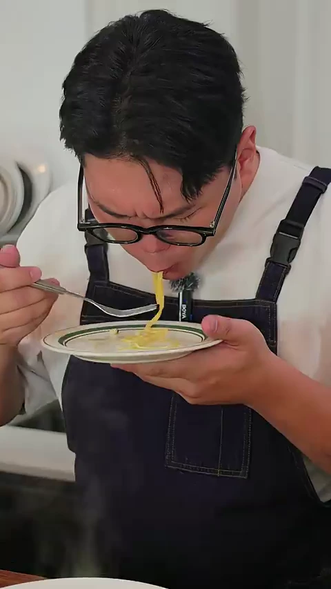 안경을 쓴 요리사가 흰색 티셔츠에 앞치마를 두르고 포크로 파스타 면을 돌돌 말아 먹고 있는 모습