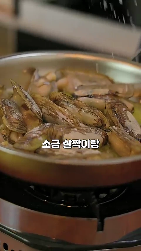 프라이팬 안에서 맛조개들이 기름과 어우러져 익어가고 있는 모습이며, 화면 중앙에 '소금 살짝이랑'이라는 자막이 떠 있습니다.