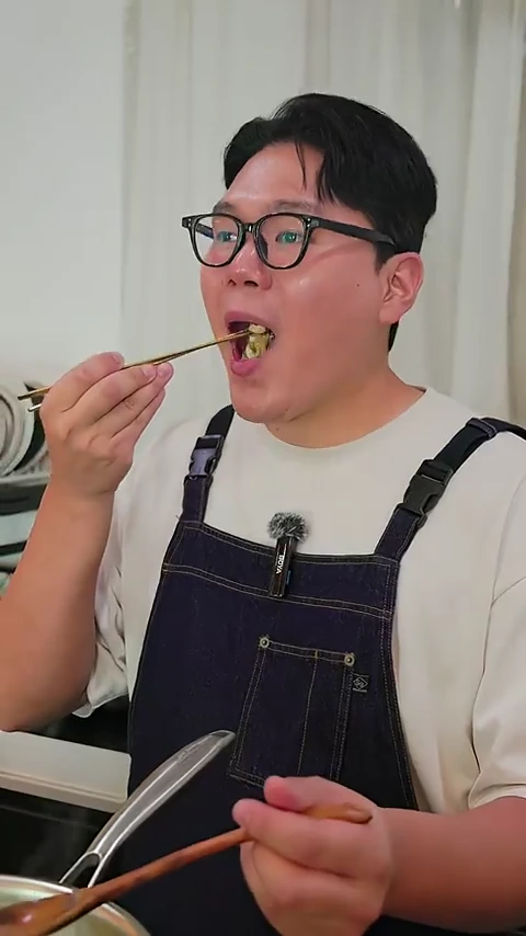 안경을 쓴 남성이 앞치마를 두르고 젓가락으로 완성된 맛조개 요리를 집어 입으로 가져가 맛보는 모습